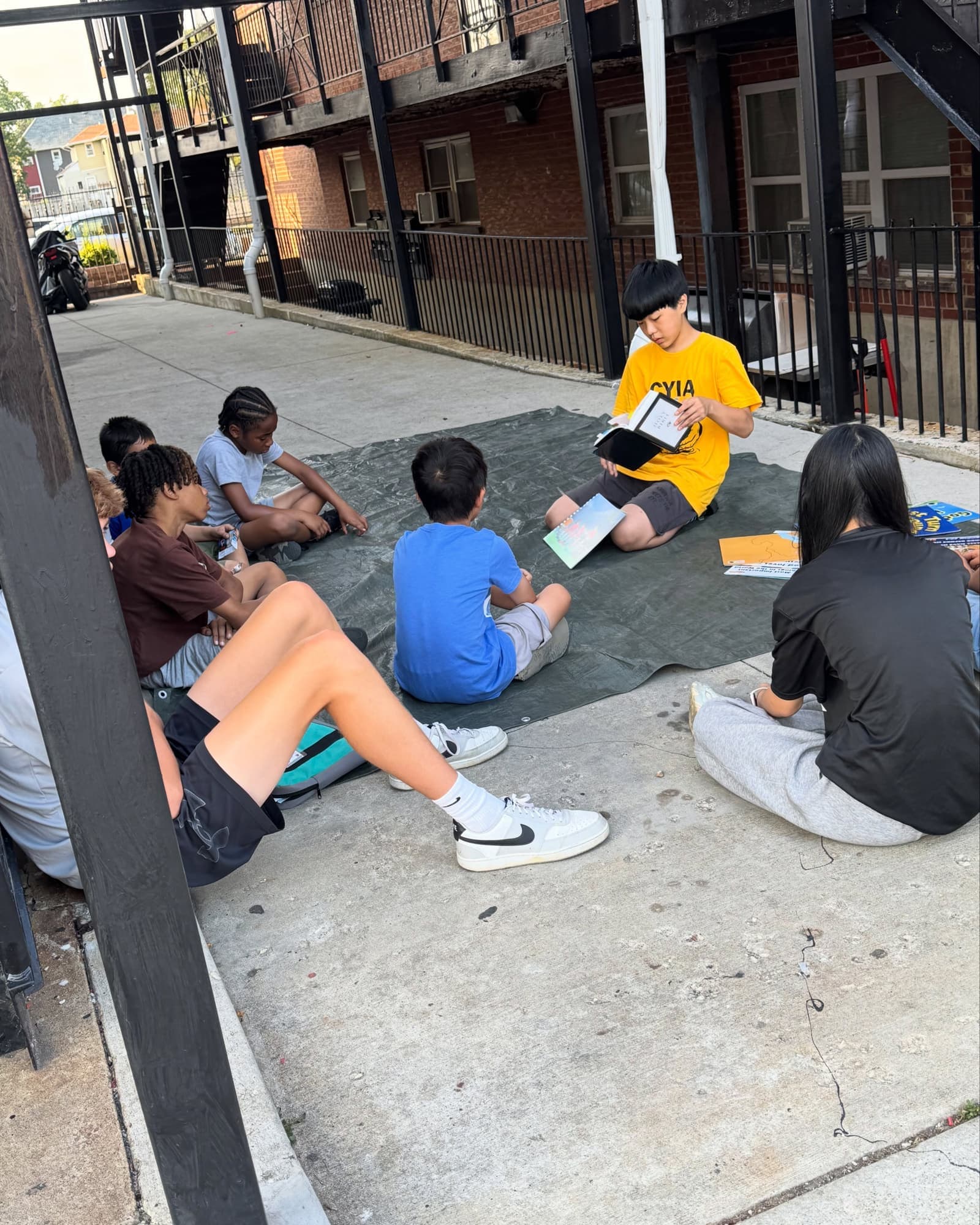 A teenager in a yellow CYIA shirt teaches children sitting on a tarp beside an apartment building