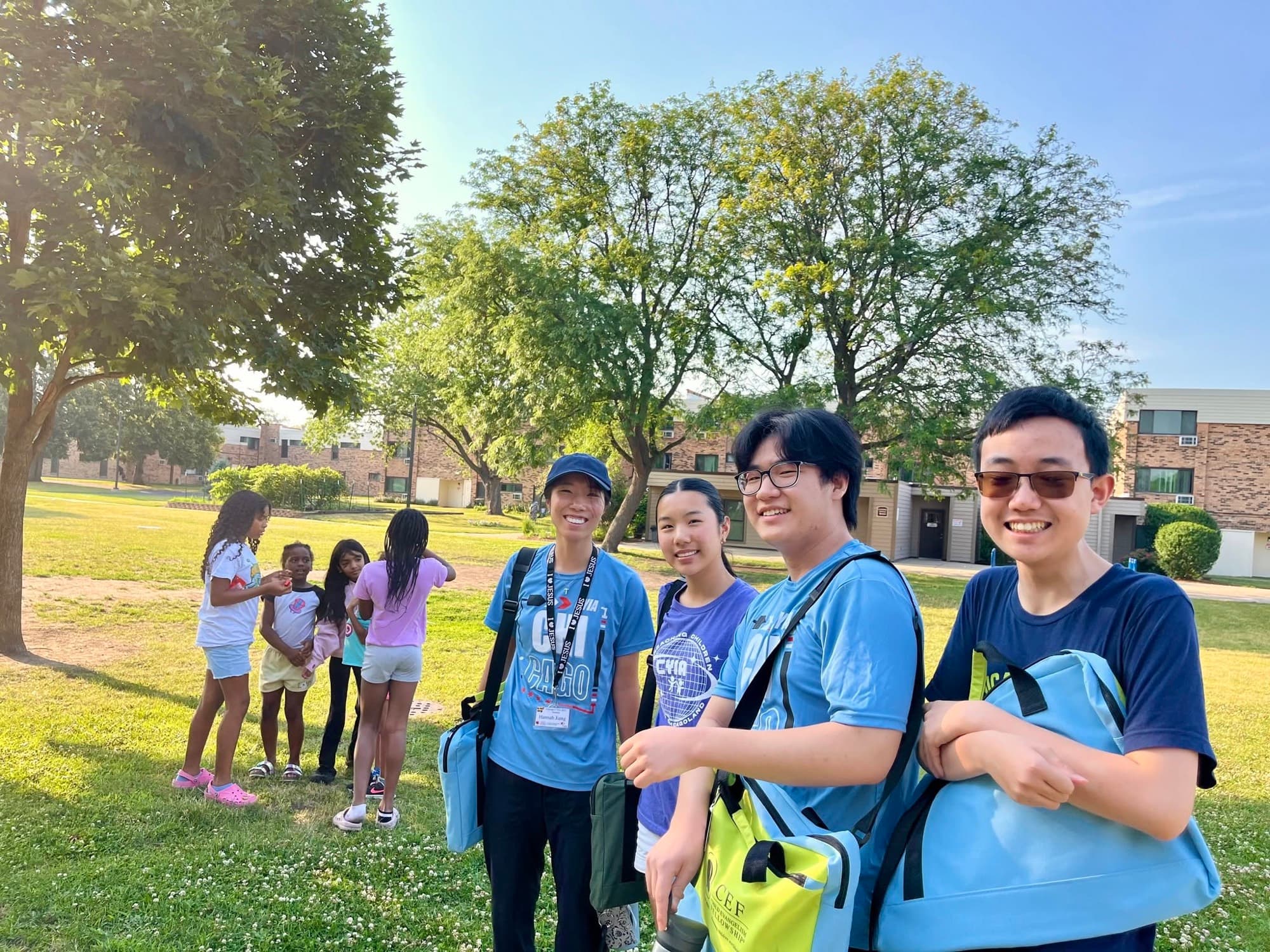 Three leaders in blue shirts smile outdoors while children gather behind them